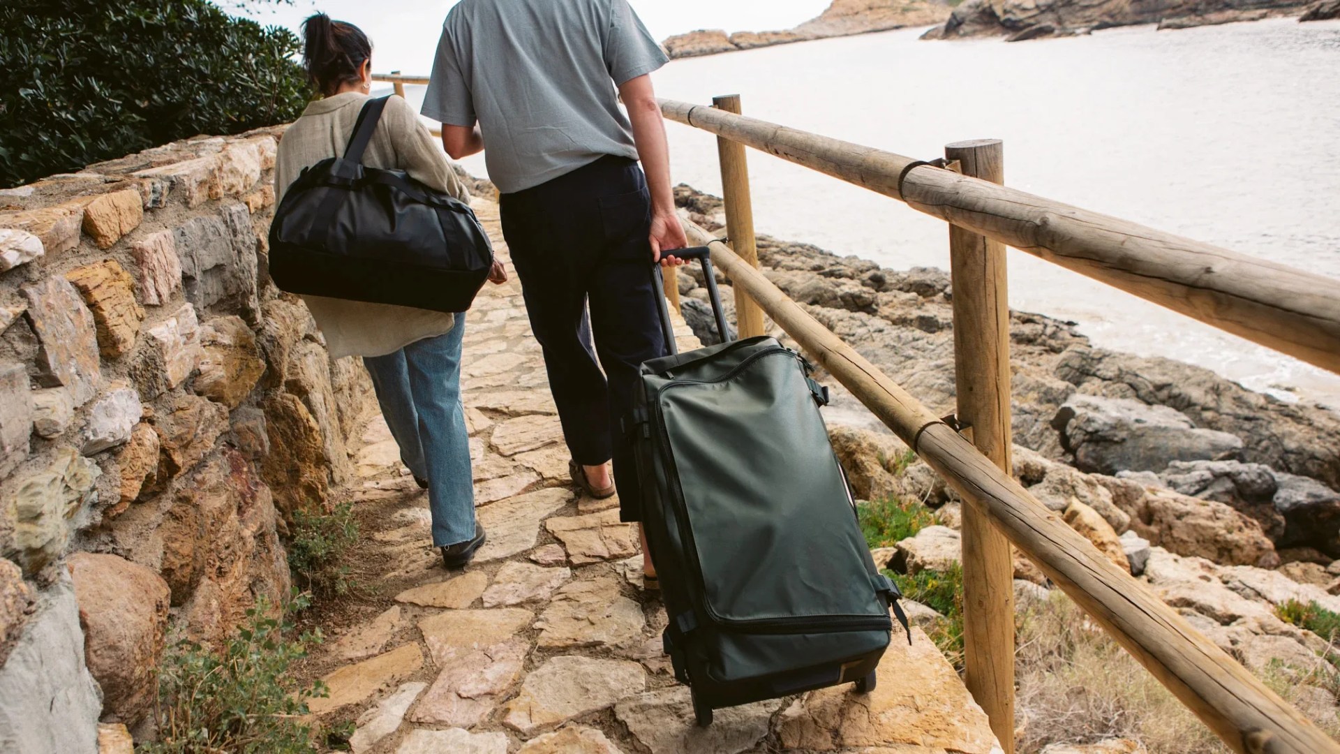 two people walking on a path near water with luggage