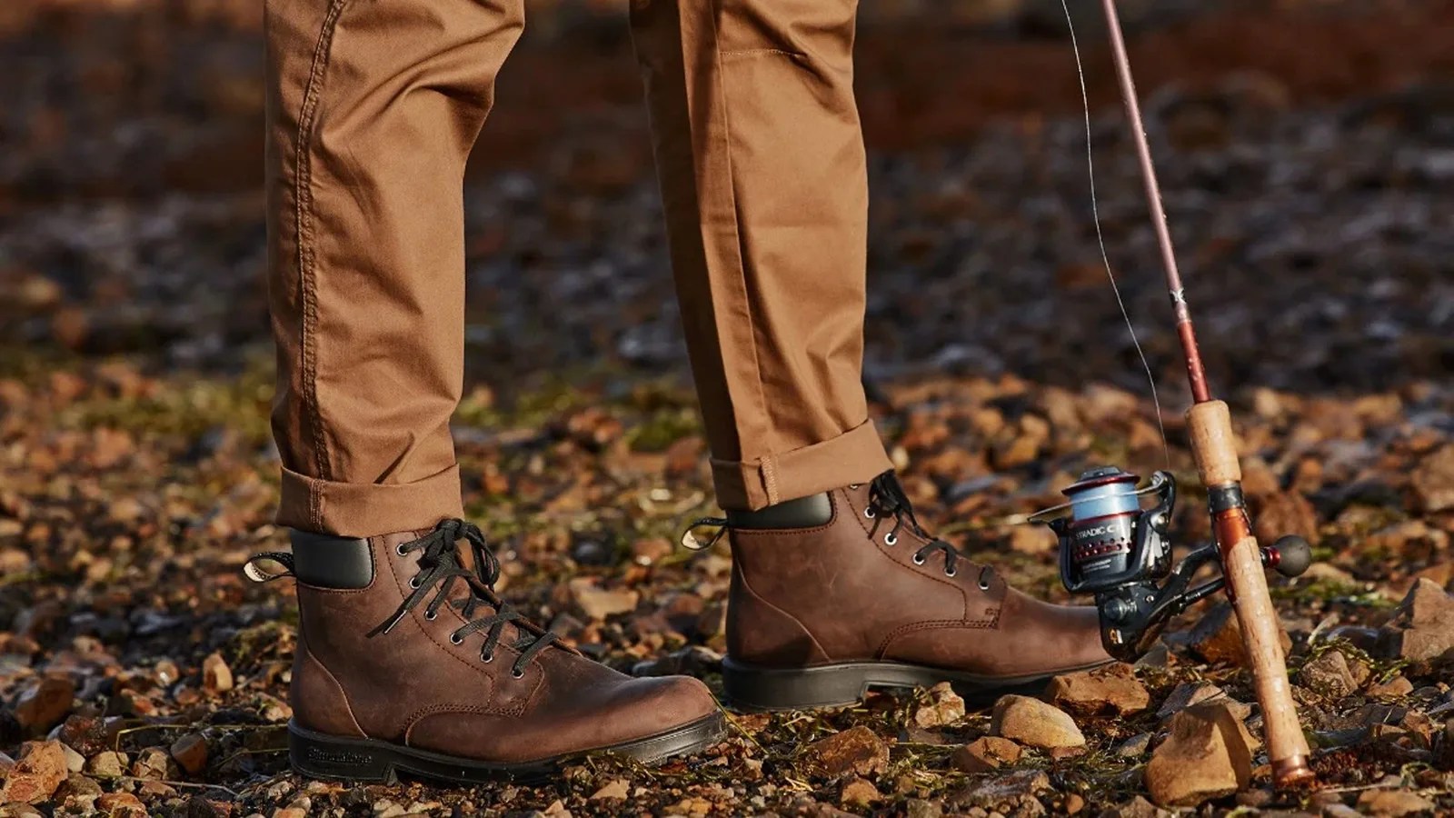 A person in khaki pants pictured below the knees wearing a pair of brown blundstone lace up boots standing on a rocky patch of ground holding a fishing pole