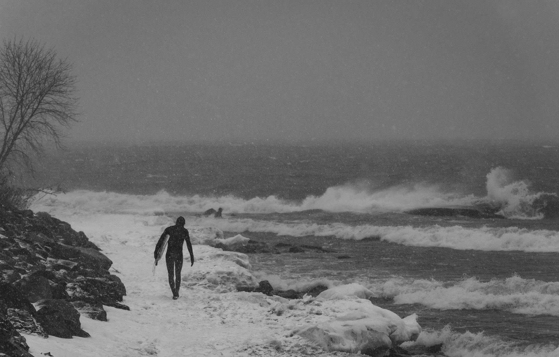 A person in a wetsuit with a surfboard walks along a frozen coastline in the snow