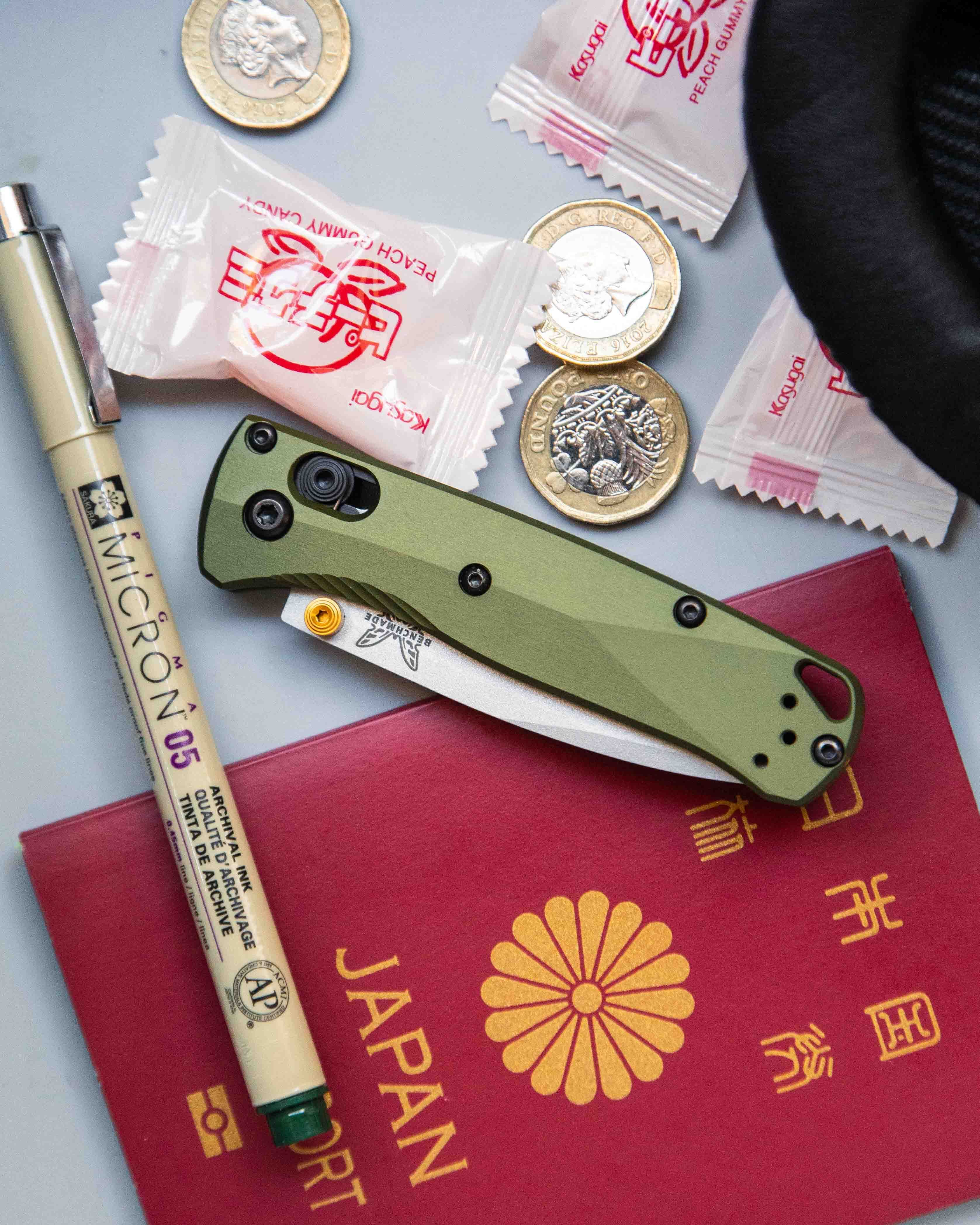 Benchmade knife surrounded by passport, pen, coins, candy and headphones on gray table